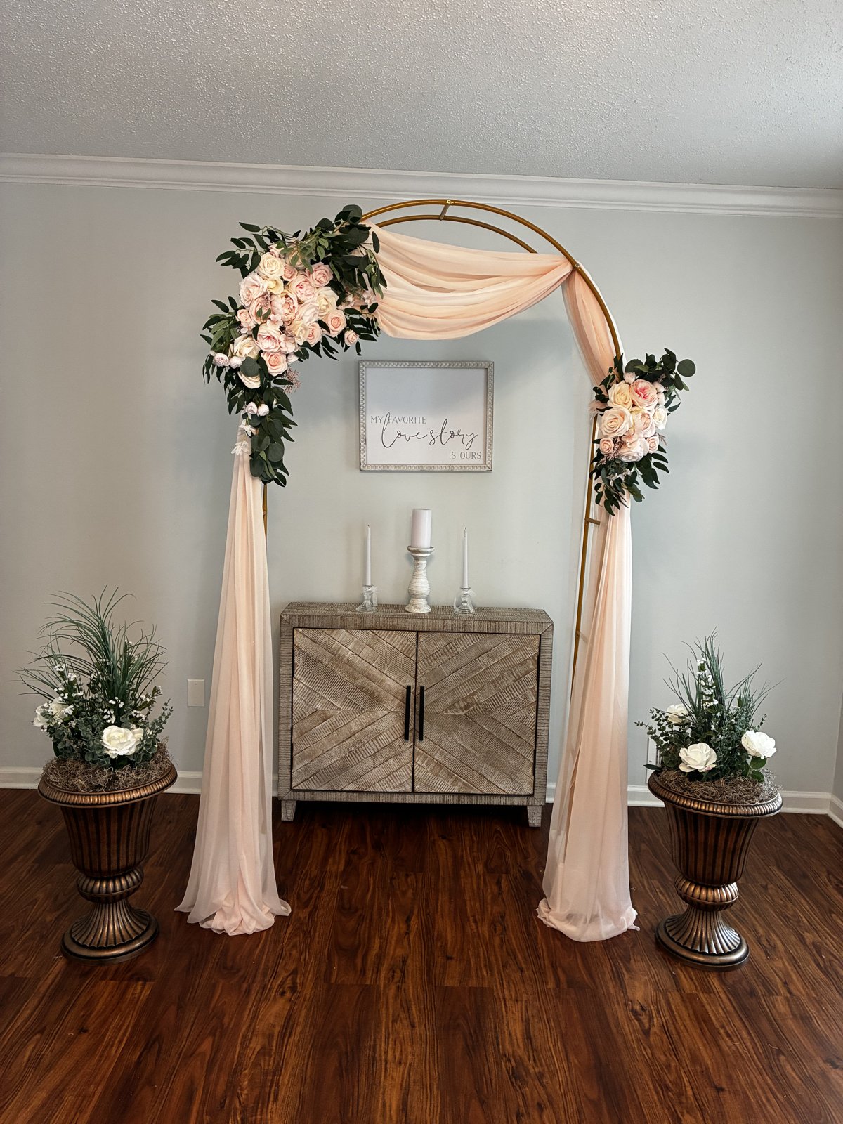 The gold ceremony arch draped in blush fabric with pink and ivory roses, a wooden cabinet with unity candles, and flower urns on each side